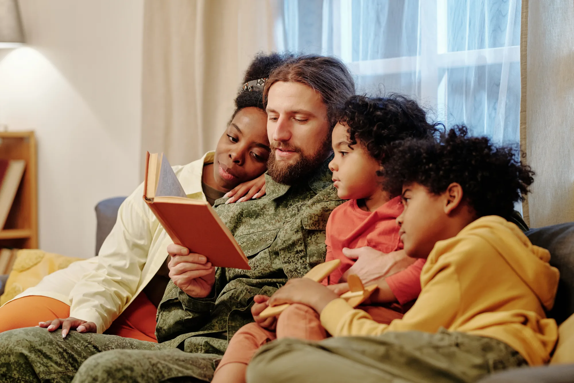 A military dad in uniform reading a storybook to his wife and two children on the couch