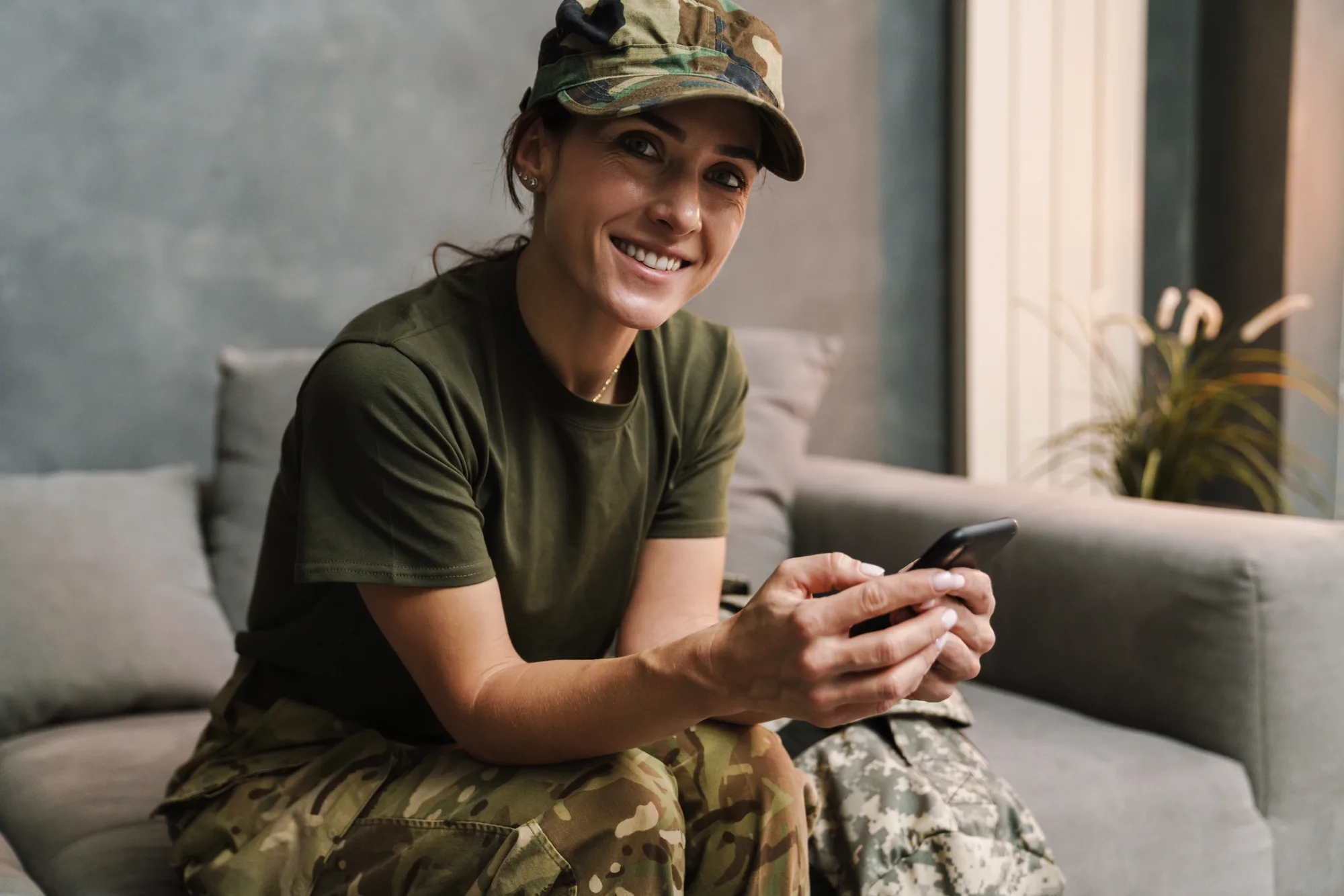 A smiling servicewoman in a camo cap holding her phone on a couch